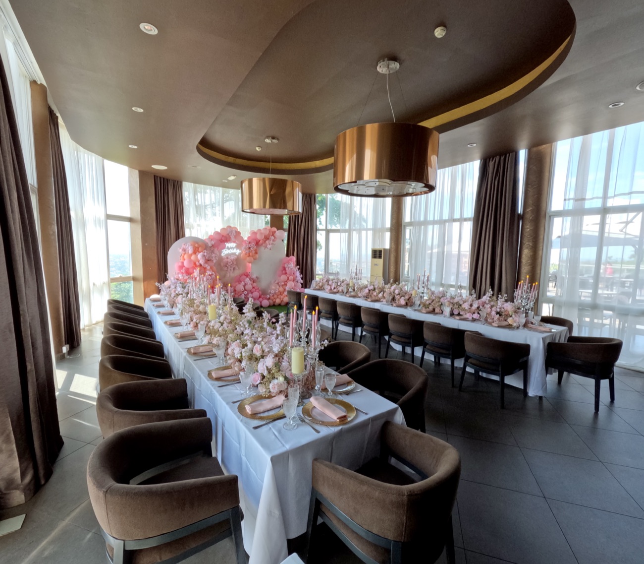 Long guest table dressed with blush floral arrangements and pink taper candles inside the VIP Room at Skyz Hotel Kampala, tall windows framing the city skyline